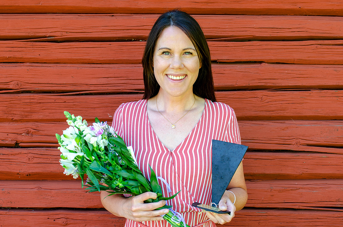 Anna Beck med blommor i höger hand och utmärkelsen Kilen i vänster hand. Hon står framför en faluröd vägg.