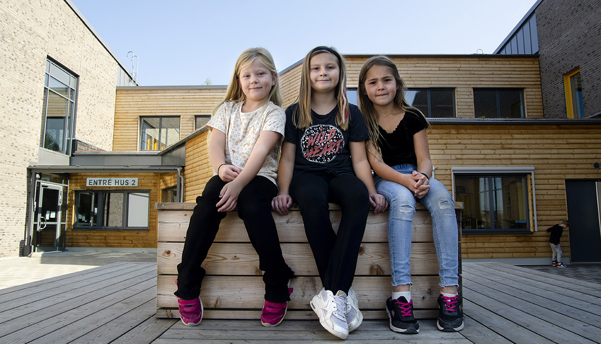 Tre flickor sitter på bänk utanför träbyggnad