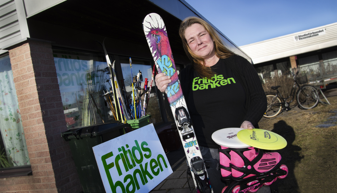 Kvinna med slalomskidor, frisbee och rullskridskor i händerna står framför byggnad. Skylt i bakgrunden med texten: Fritidsbanken.